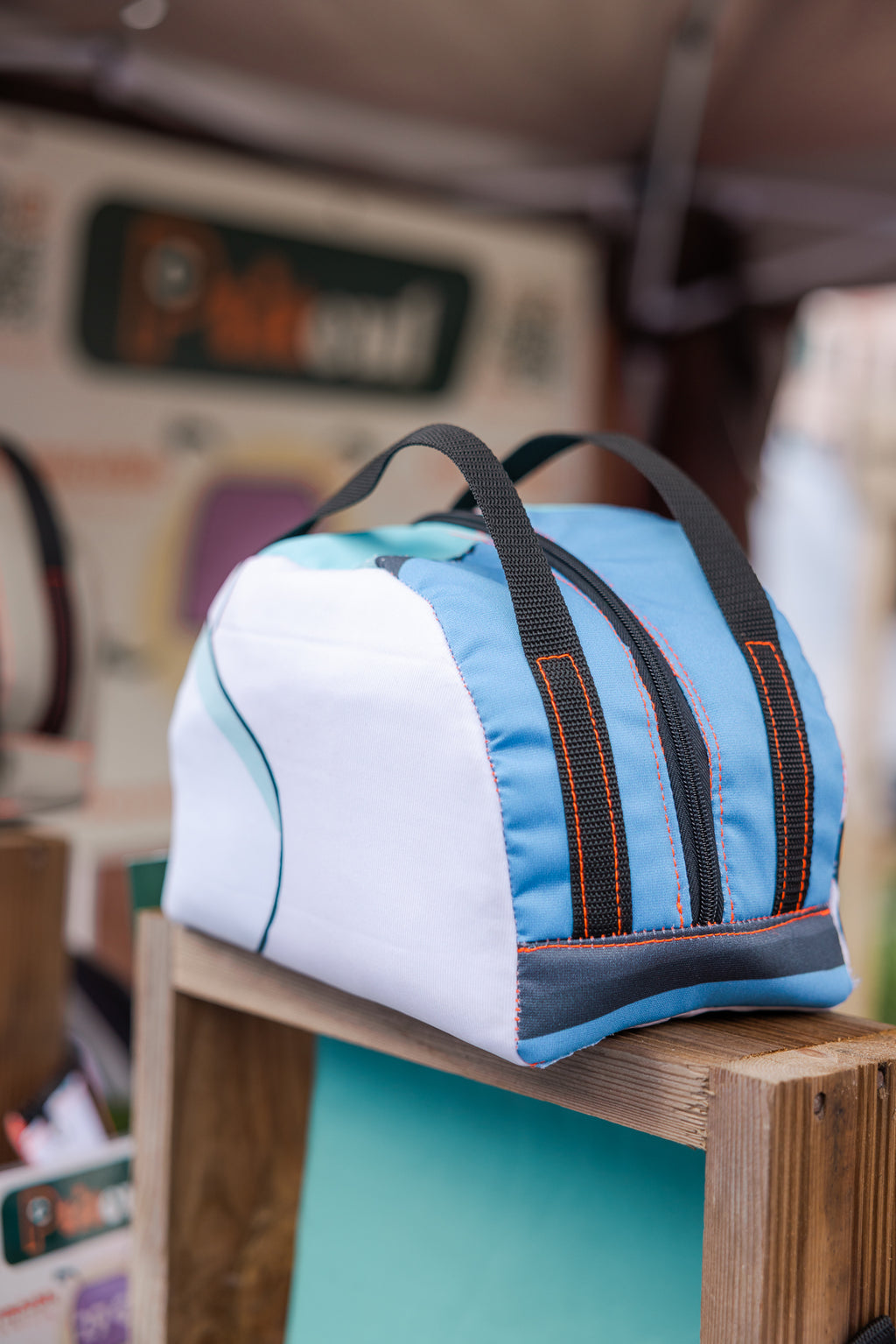 Sac en tissu épais, avec une fermeture éclair sur le dessus. Il est principalement blanc et bleu clair avec des coutures oranges et deux poignées marrons.